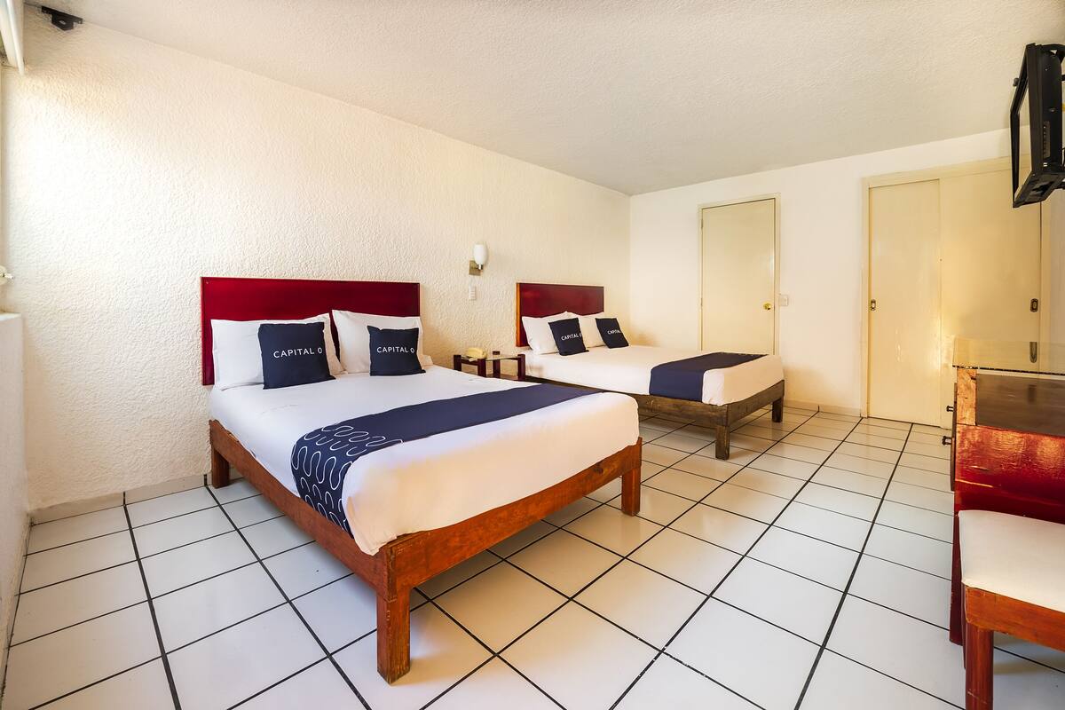 A hotel room with two beds, each with a red headboard and white bedding. There is a small table and a chair in the corner.