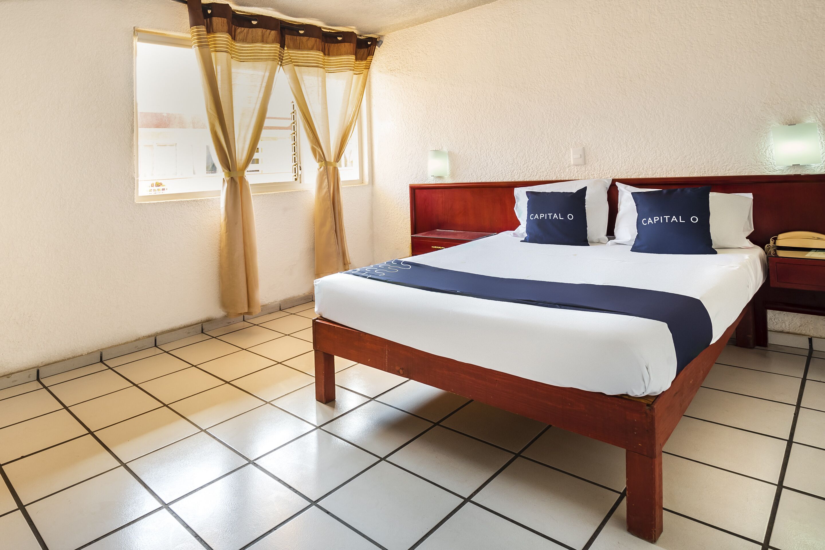 A hotel room with a bed, a nightstand, a lamp, and a window with curtains.