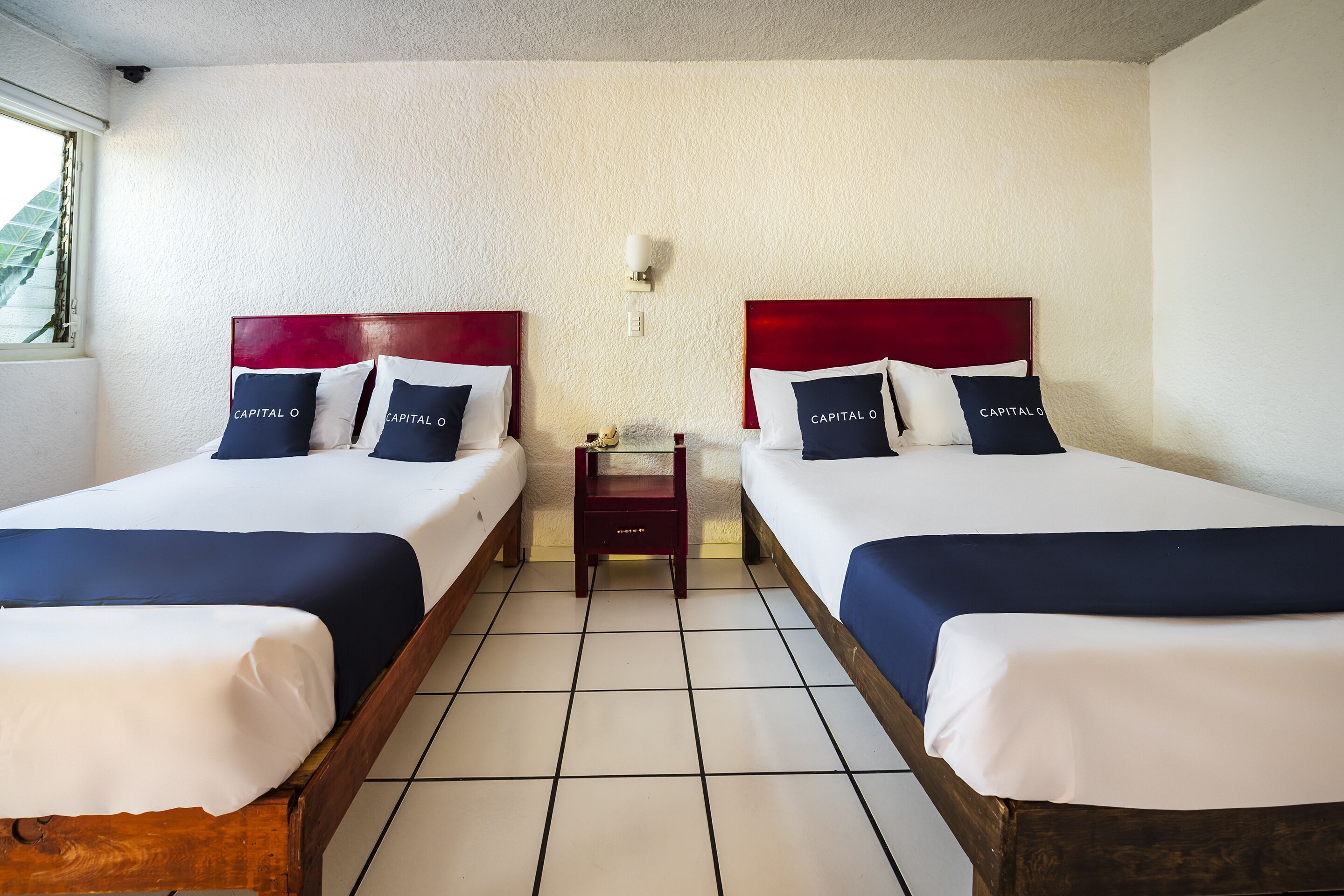 A hotel room with two beds, each with a red headboard and white bedding. There is a small wooden nightstand between the beds with a lamp and a small table. The room has white walls and a tiled floor.