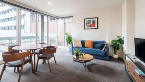 A modern living room with a sofa, coffee table, and chairs, a TV, and large windows with a view of buildings.