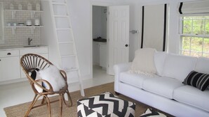 A modern kitchen with white cabinetry, a white countertop, and a built-in shelf. There is a white chair with a white cushion, a white sofa with a black and white patterned throw, and a window with a white curtain. - Beautiful Family Home Nestled In The Heart Of Sagaponack (Sagaponack)