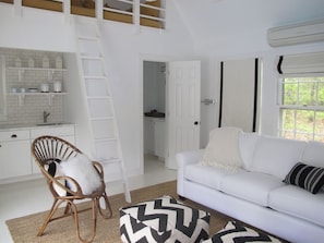 A modern kitchen with white cabinetry, a white countertop, and a built-in shelf. There is a white chair with a white cushion, a white sofa with a black and white patterned throw, and a window with a white curtain.