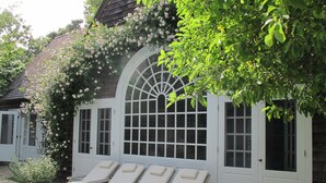 A covered outdoor seating area with lounge chairs and a large arched window. - Beautiful Family Home Nestled In The Heart Of Sagaponack (Sagaponack)