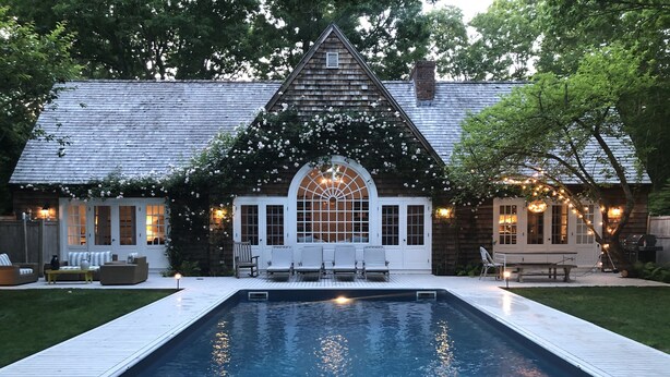 A pool with a wooden deck, a covered seating area with white chairs, and a building with a gabled roof and large windows. - Beautiful Family Home Nestled In The Heart Of Sagaponack (Sagaponack)