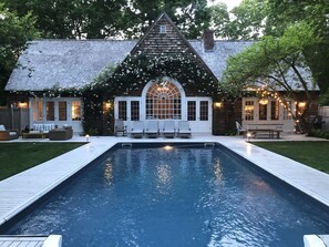 A pool with a wooden deck, a covered seating area with white chairs, and a building with a gabled roof and large windows. - Beautiful Family Home Nestled In The Heart Of Sagaponack (Sagaponack)