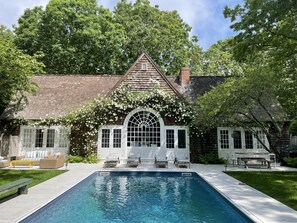A poolside view of a house with a wooden exterior, white-framed windows, and a covered patio area with outdoor furniture. - Beautiful Family Home Nestled In The Heart Of Sagaponack (Sagaponack)