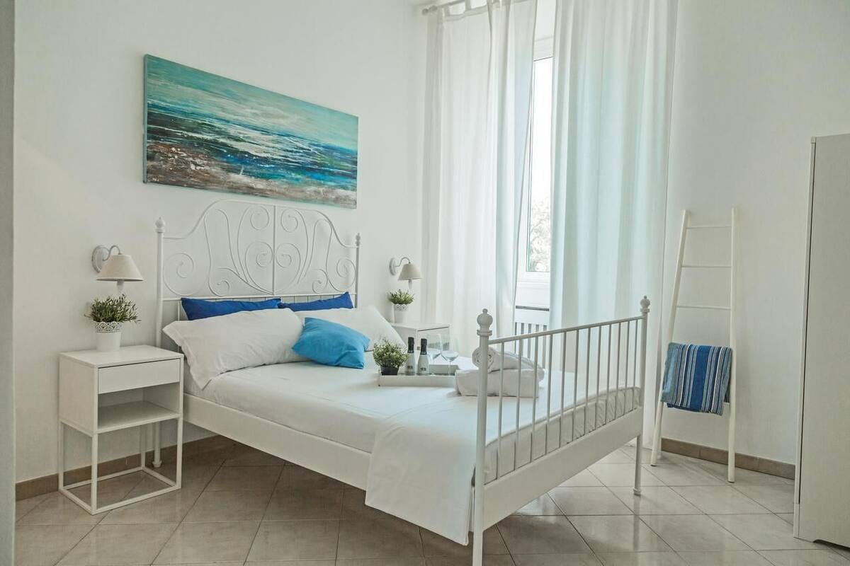 A white four-poster bed with a headboard, two bedside tables with lamps, a painting of a seascape, a window with sheer curtains, and a white wardrobe.