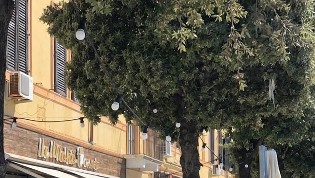 Un cafè a l'aire lliure amb taules i bancs de fusta, envoltat d'edificis amb balcons i tendals.