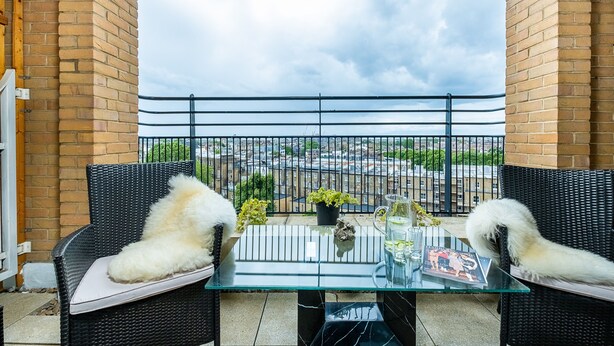 A balcony with wicker furniture, a glass table, and a view of a cityscape.