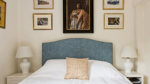 Un lit bien fait, avec une tête de lit bleue, une literie blanche et un seul coussin décoratif. Des tableaux encadrés sont accrochés au mur au-dessus du lit.