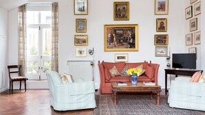Un salon avec un canapé rouge, deux fauteuils, une table basse et un téléviseur. Les murs sont ornés de tableaux encadrés et un lustre est suspendu au plafond.