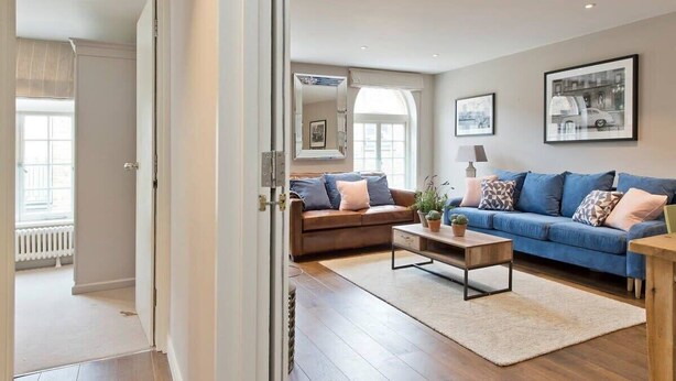 Un salon moderne avec un canapé marron, un canapé bleu et une table basse en bois. Deux tableaux encadrés ornent le mur, et une fenêtre est équipée de stores.