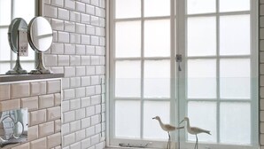 Une salle de bain aux murs recouverts de carreaux blancs, avec une grande fenêtre à vitres dépolies et deux statuettes d’oiseaux décoratives.