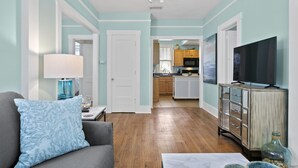 A living room with a sofa, a television, and a kitchen in the background. - Quaint Bungalow just off Main Street! (Dunedin)