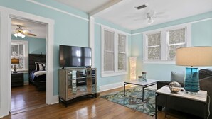 A living room with hardwood floors, a TV on a cabinet, a sofa, a coffee table, and a lamp. - Quaint Bungalow just off Main Street! (Dunedin)