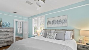 A bedroom with a large bed, a wooden dresser, a nightstand with a lamp, and a wall-mounted abstract painting. - Quaint Bungalow just off Main Street! (Dunedin)