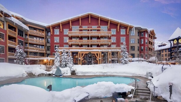 Un hotel con piscina rodeado de nieve, balcones de madera y un edificio grande al fondo.