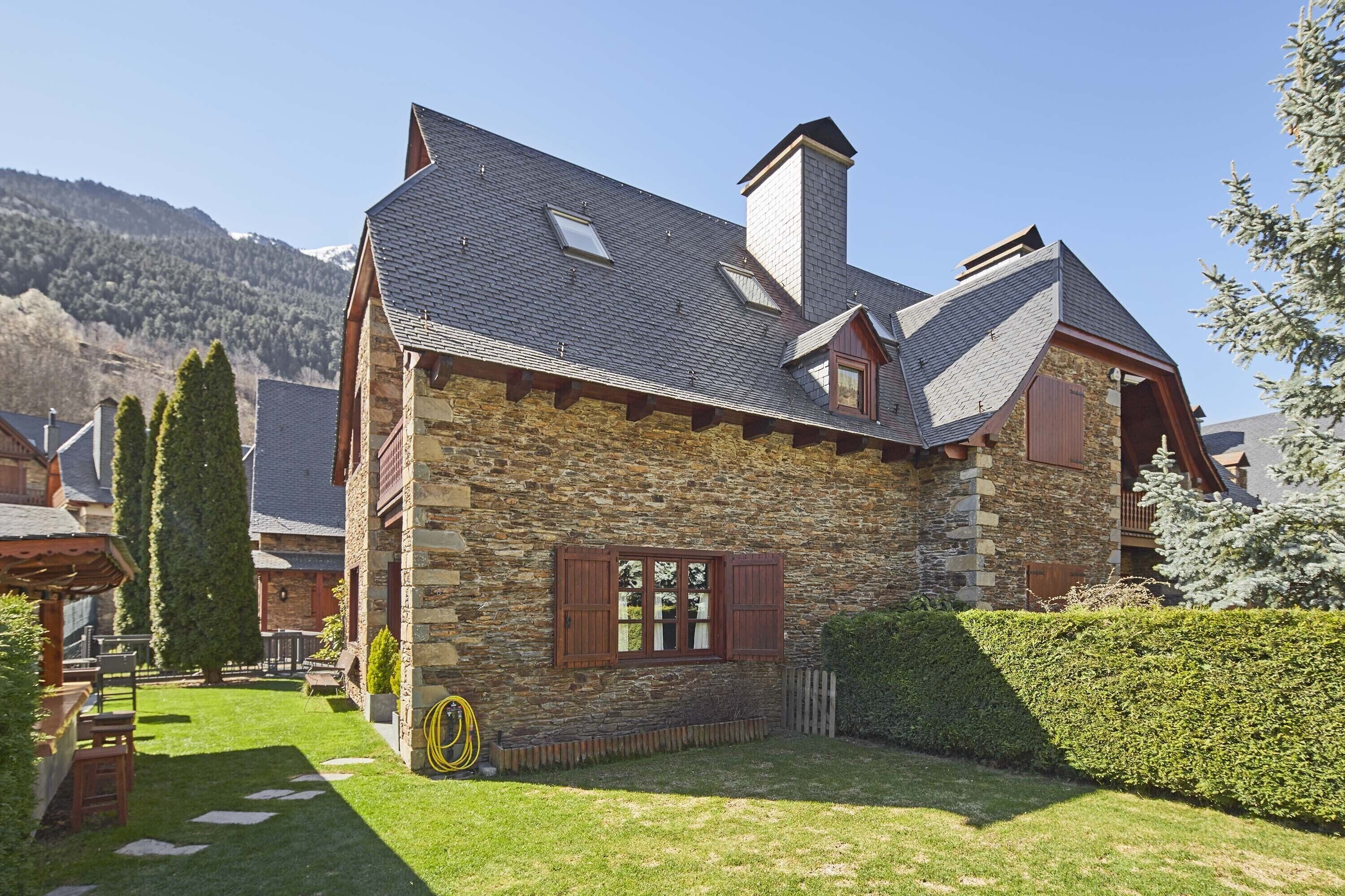 A stone house with a sloped roof, a chimney, and a small garden with a lawn and a wooden bench.