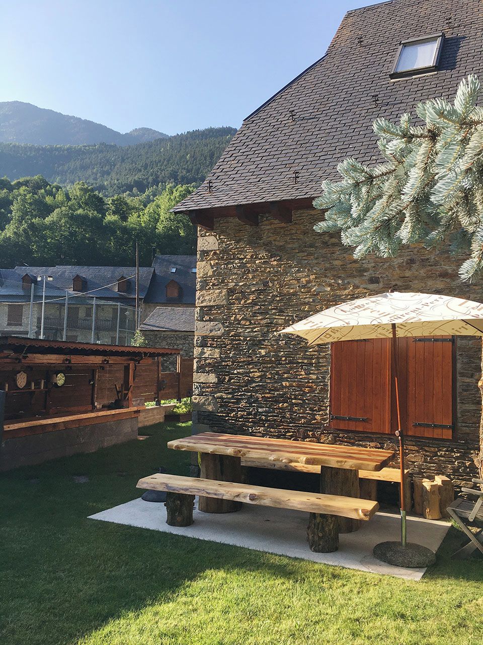A stone house with a stone wall and a wooden roof. There is a wooden bench and an umbrella in the yard.