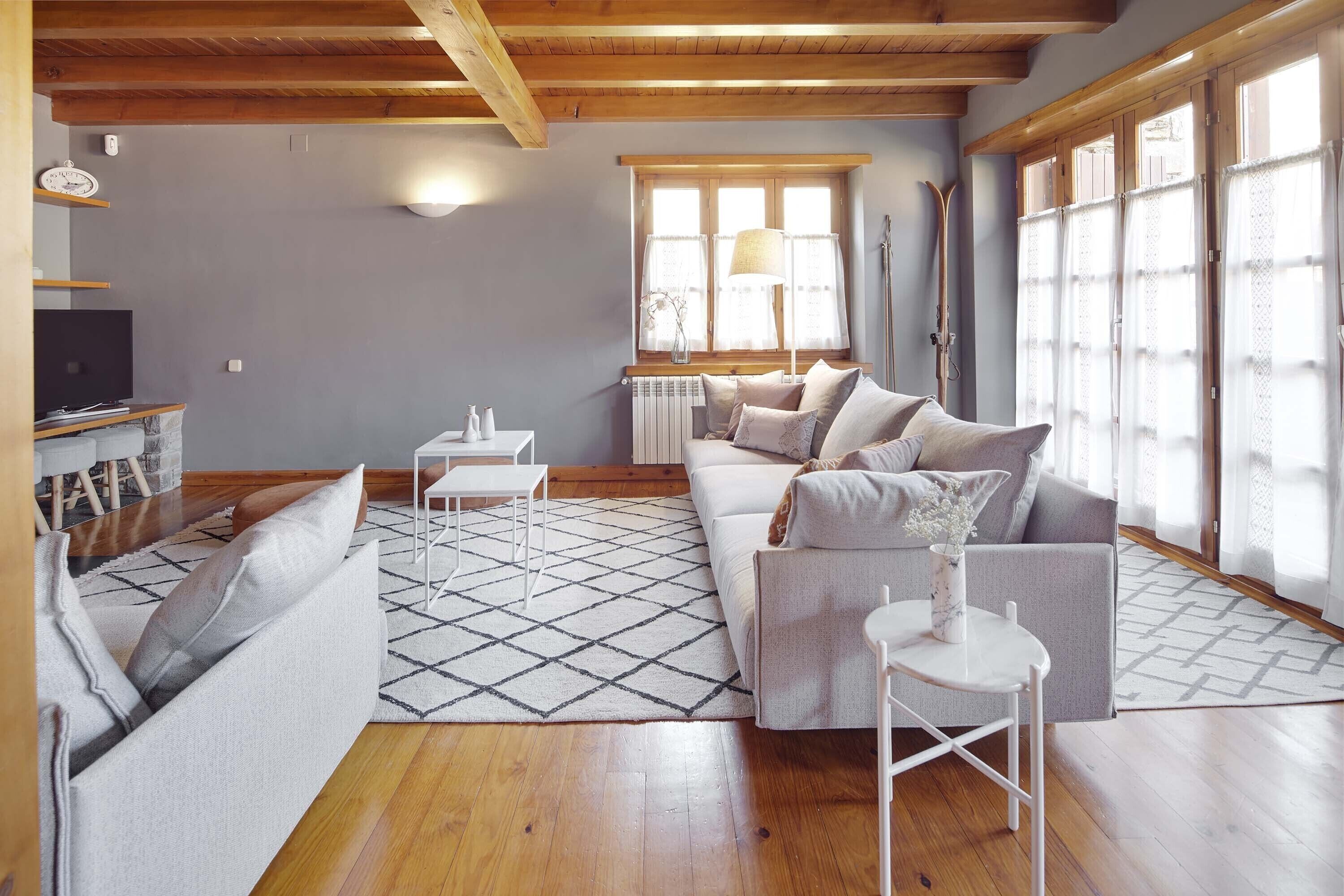 A modern living room with a wooden ceiling, a sofa set, a coffee table, and a large window with curtains.