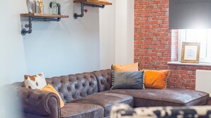 Ein braunes, ledernes Ecksofa mit orangefarbenen und grauen Kissen, eine Ziegelwand und ein an der Wand befestigter Fernseher.