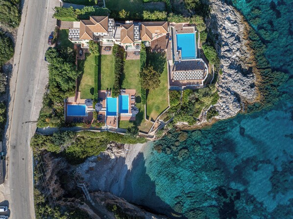 Vue aérienne d’une propriété située sur la côte, avec plusieurs piscines, un grand bâtiment doté d’un toit à motifs géométriques et une côte rocheuse.