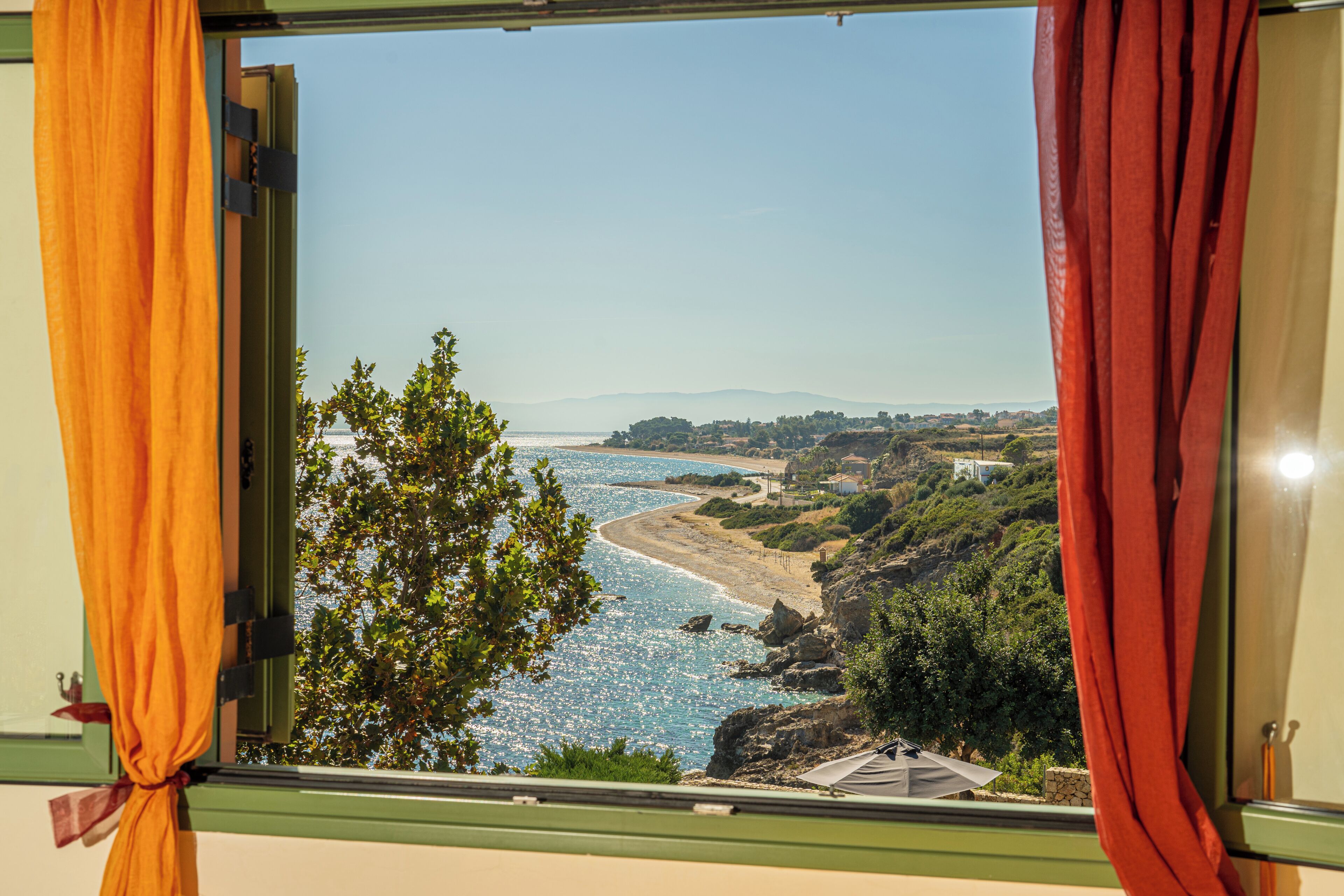 Une vue par la fenêtre sur un paysage côtier, avec un ciel dégagé, une plage et de la végétation.