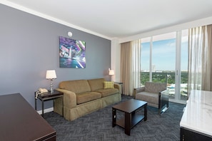 A hotel room with a gray wall, a large window with curtains, a sofa, a chair, a small table, and a painting.