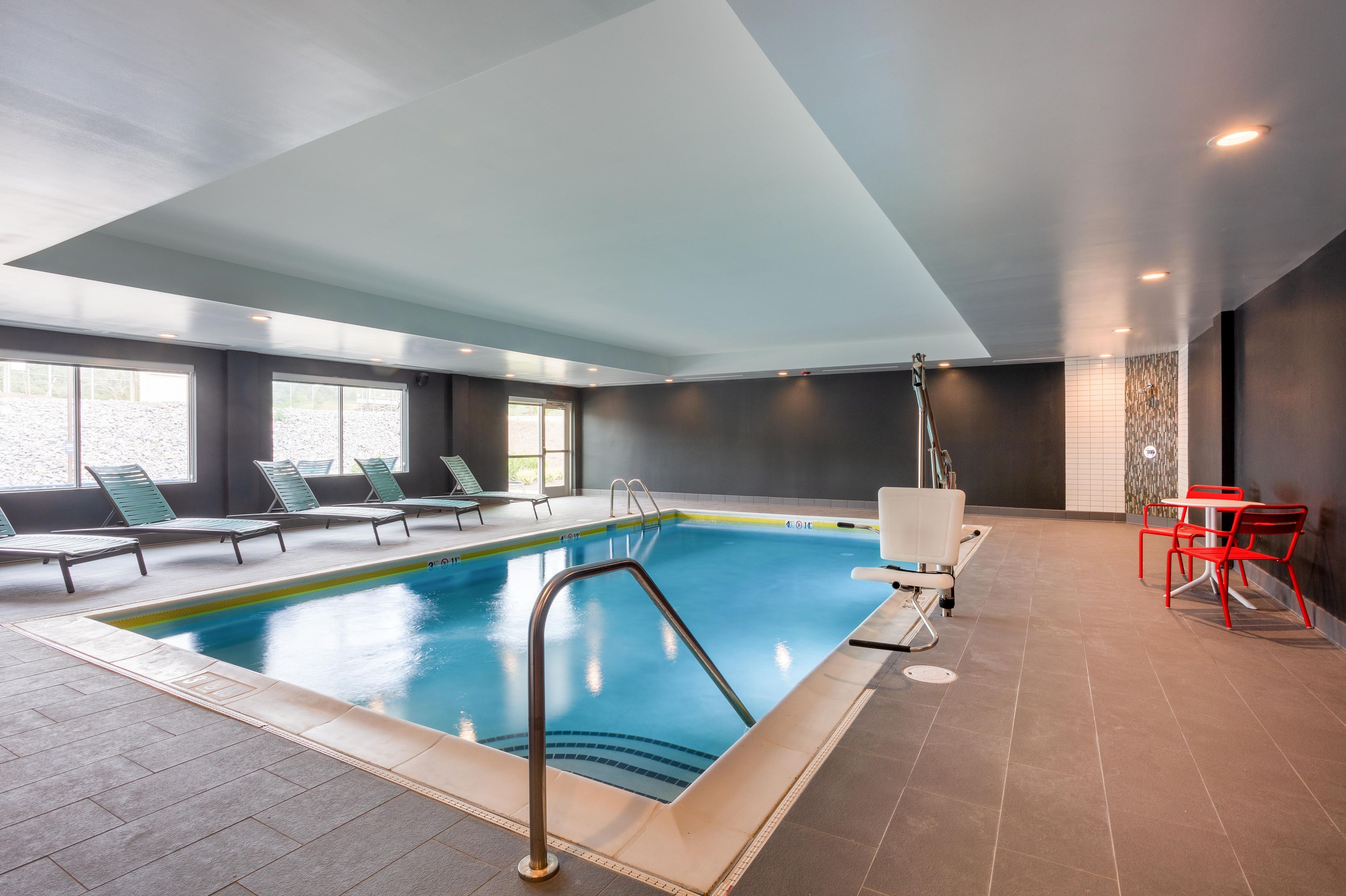 An indoor swimming pool with a clear blue water, surrounded by lounge chairs and a small table with chairs.