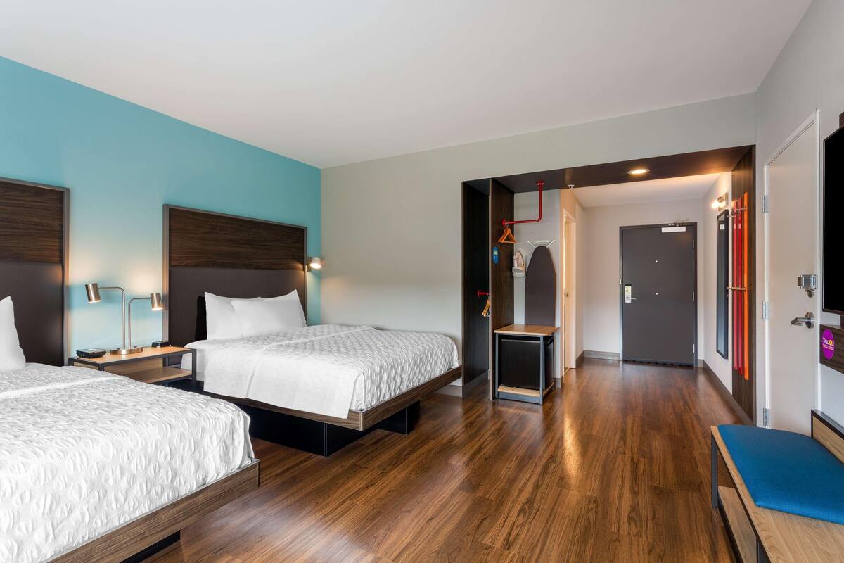 Habitación de hotel con dos camas, piso de madera, baño con espejo y perchero, y una puerta que da a otra habitación.