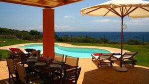 A poolside area with a table set for dining, lounge chairs, and a large umbrella. - Villa A, Brio Exclusive | 3BR Seafront Family Villa with Private Pool & Jetty (Kefalonia Islands)