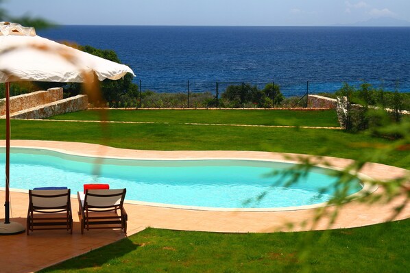 A swimming pool with two lounge chairs and a white umbrella, surrounded by a well-maintained lawn and a view of the sea.