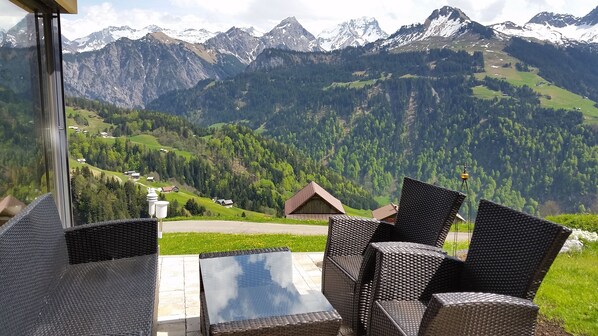 Terraço com móveis de vime e uma mesa de vidro, com vista para uma paisagem montanhosa com casas e vegetação.