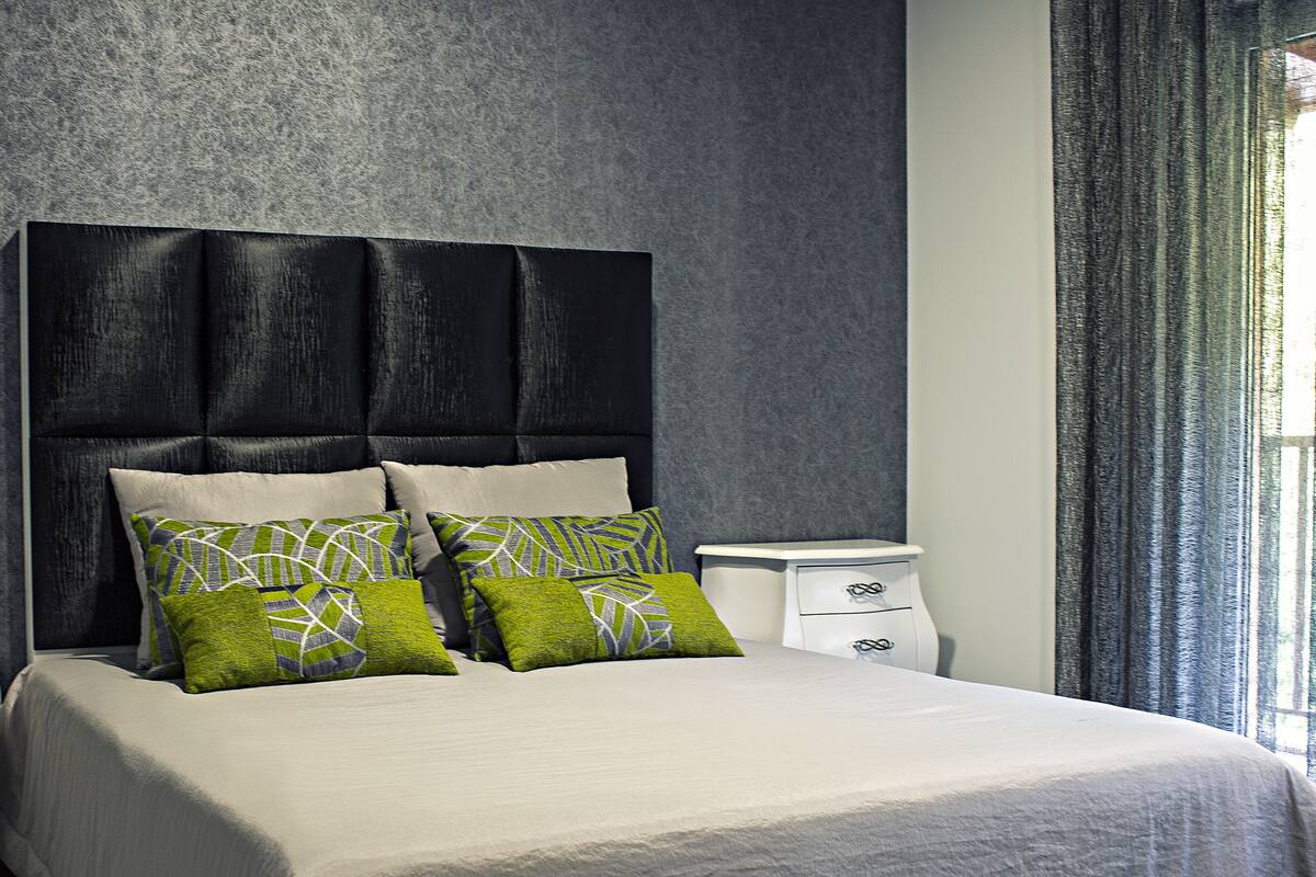 A neatly made bed with a large headboard, two bedside tables, and a window with curtains.