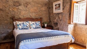 A bedroom with a wooden bed, stone walls, a small bedside table with a plant, and a window with a wooden frame. - Casa Moinho da Mouta (Sabrosa)