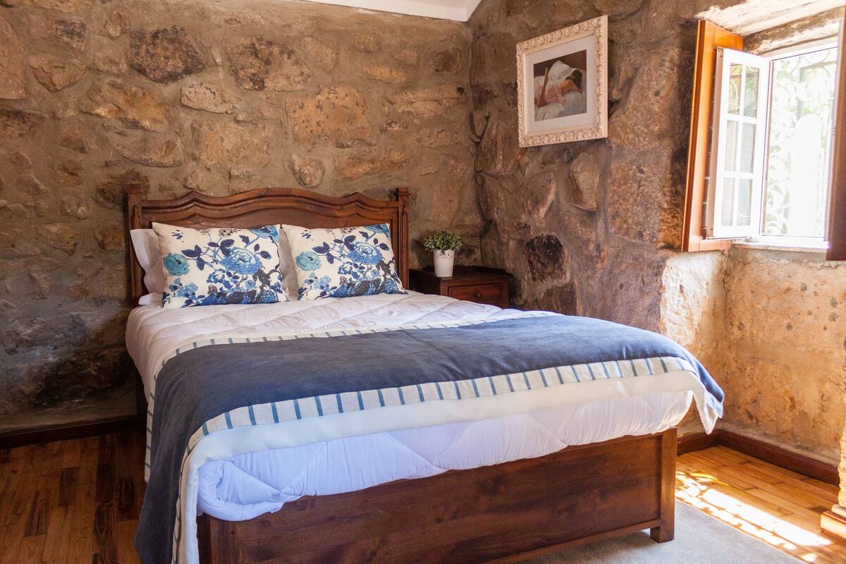A bedroom with a wooden bed, stone walls, a small bedside table with a plant, and a window with a wooden frame.