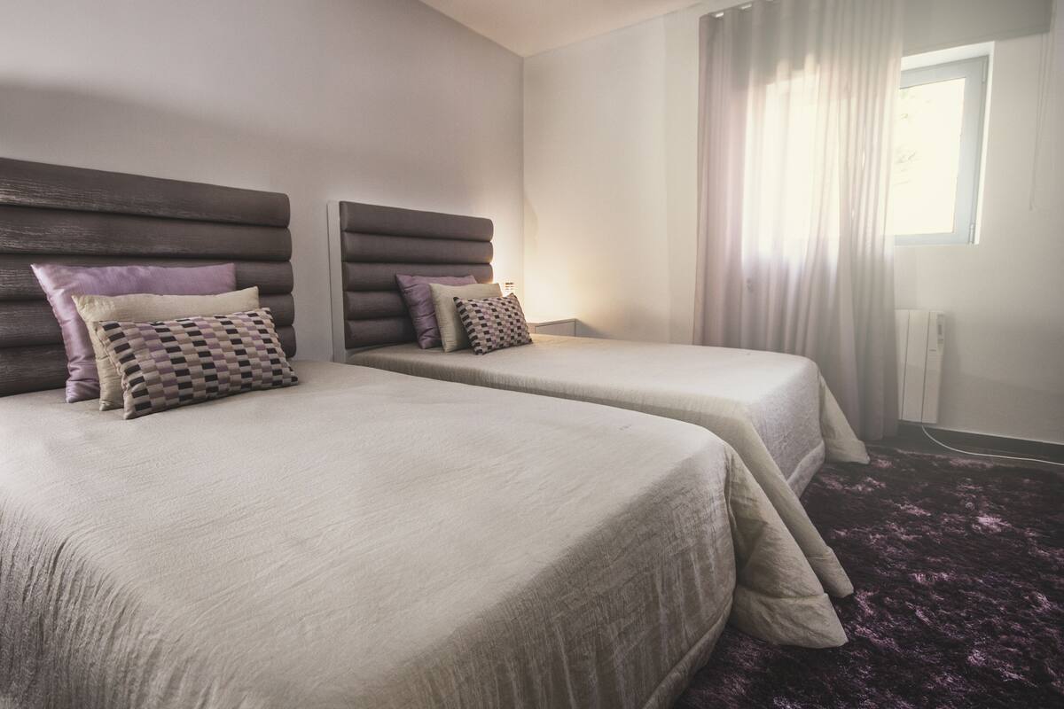 A hotel room with two beds, each with a headboard, a window with curtains, and a purple carpet.