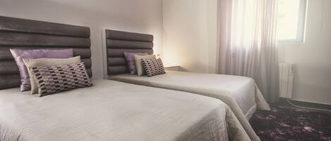 A hotel room with two beds, each with a headboard, a window with curtains, and a purple carpet.