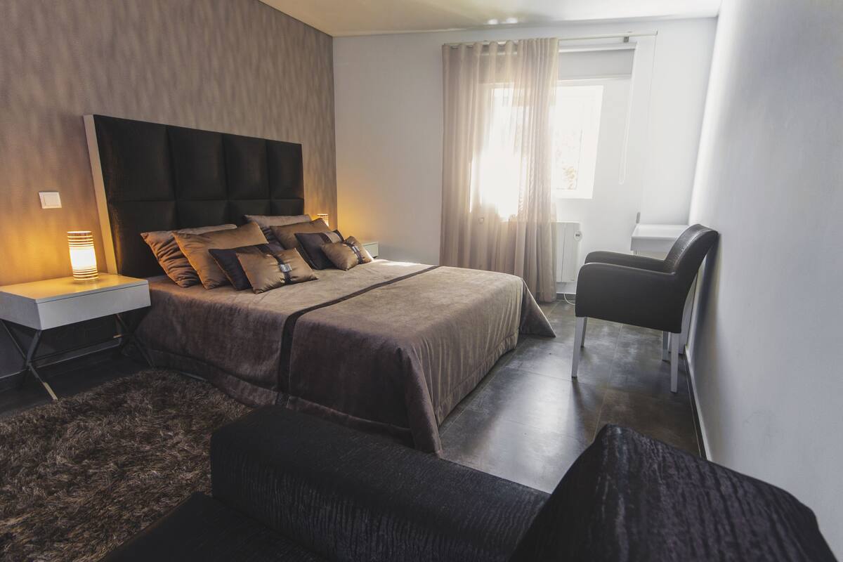 A modern hotel room with a large bed, a desk with a lamp, a chair, and a TV mounted on the wall.