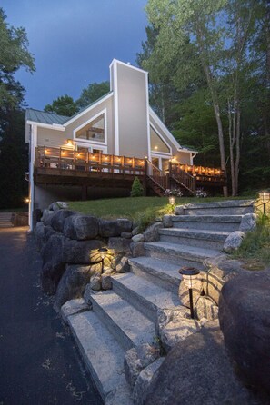 A modern house with a stone staircase leading to a wooden deck, surrounded by a stone wall and a well-maintained lawn.