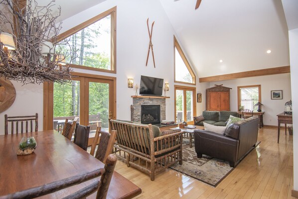 A spacious living room with a fireplace, wooden furniture, and large windows offering a view of the outdoors.