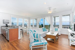 A spacious living area with a kitchen, white furniture, and large windows offering a view of the ocean. - SC202 Coastal LegaSea by First Flight Rentals (Kill Devil Hills)