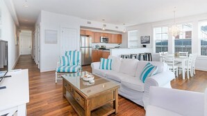 Een ruime woonkamer met een witte bank, gestreepte fauteuils, een houten salontafel en een keuken op de achtergrond.