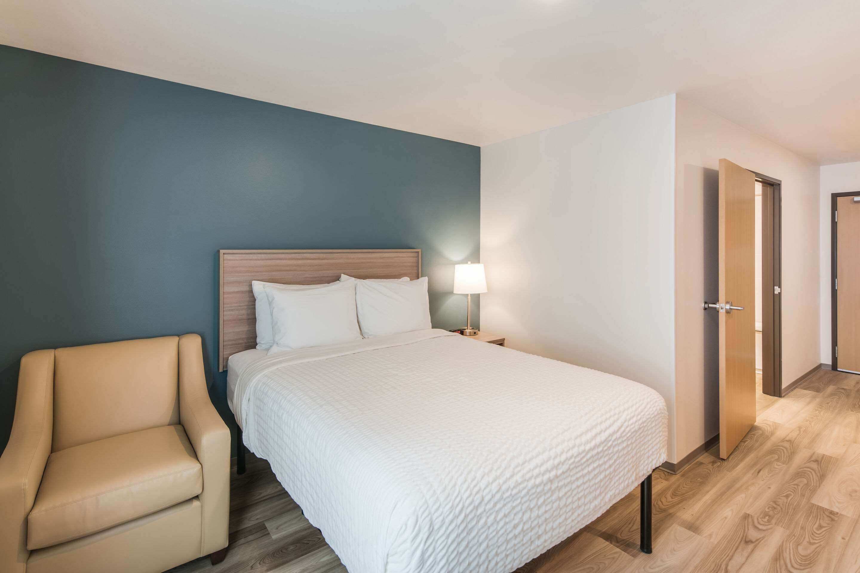 Una habitación de hotel con una cama, una silla, una mesita de noche con lámpara y una puerta.