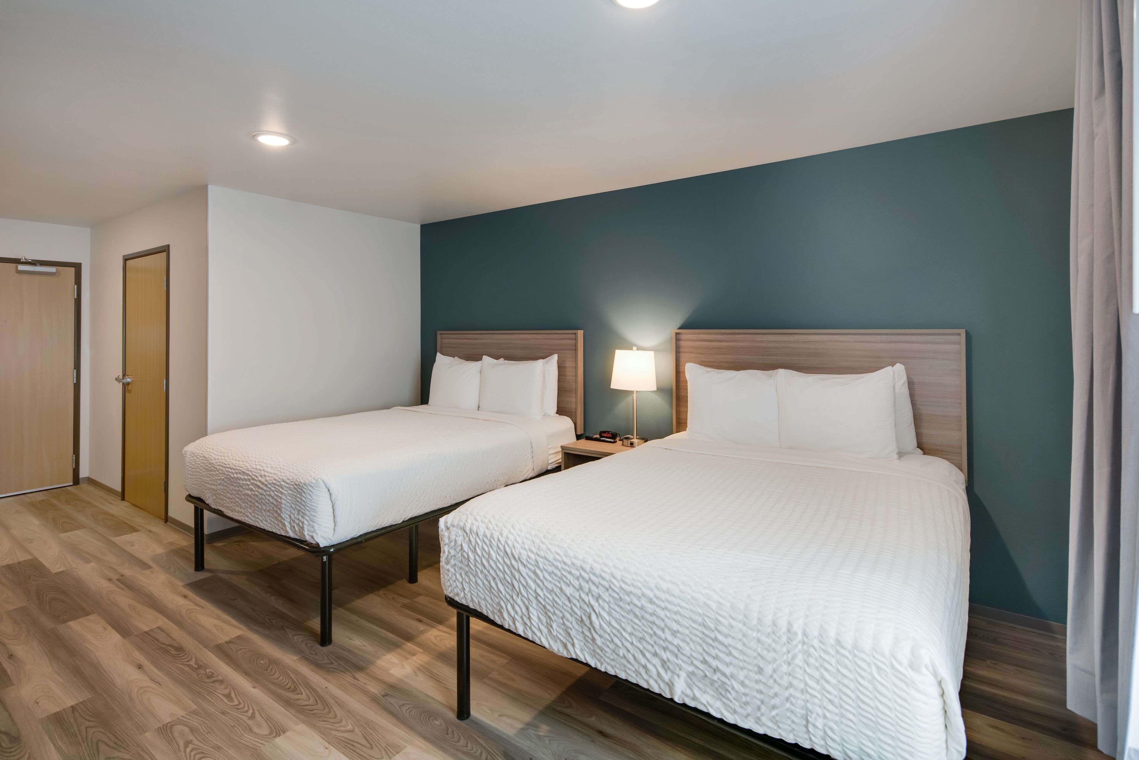 A hotel room with two beds, a nightstand with a lamp, wooden flooring, and a teal accent wall.
