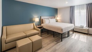 A hotel room with a bed, a sofa, a nightstand, and a window with curtains. - WoodSpring Suites Tri-Cities - Richland  (Richland)