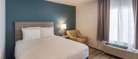 Quarto de hotel com cama, cadeira, mesa de cabeceira, luminária e janela com cortinas.