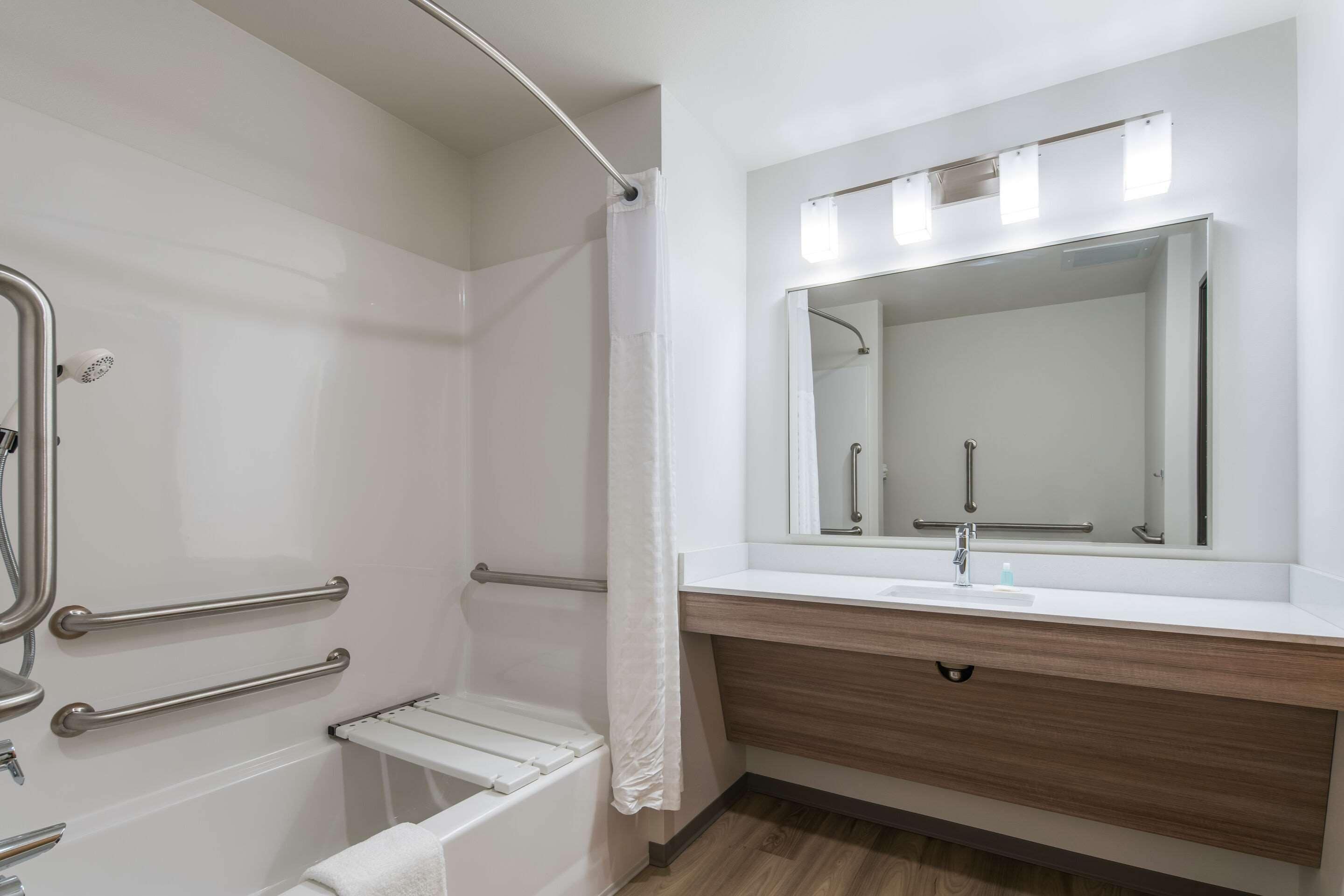 A modern bathroom with a shower, grab bars, a white sink, and a mirror with lights.