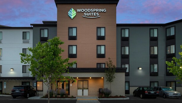 A multi-story hotel with a prominent "WOODS SPRING SUITES" sign, featuring a modern design with a mix of brick and panel exteriors. - WoodSpring Suites Tri-Cities - Richland  (Richland)