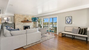 A spacious living room with a stone fireplace, a large sofa, a bench, and a view of the outdoors through large sliding glass doors. - Oceanfront dining, shopping, Pool, spa, and sauna on site!- The Harbor Penthouse (Oceanside)